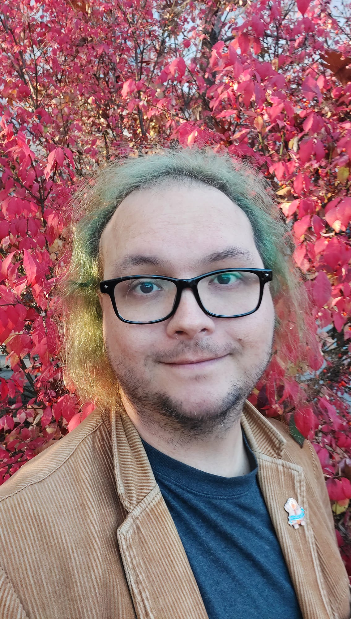 A man with green hair wearing a brown corderoy jacket stands in front of a bright red bush looking happy